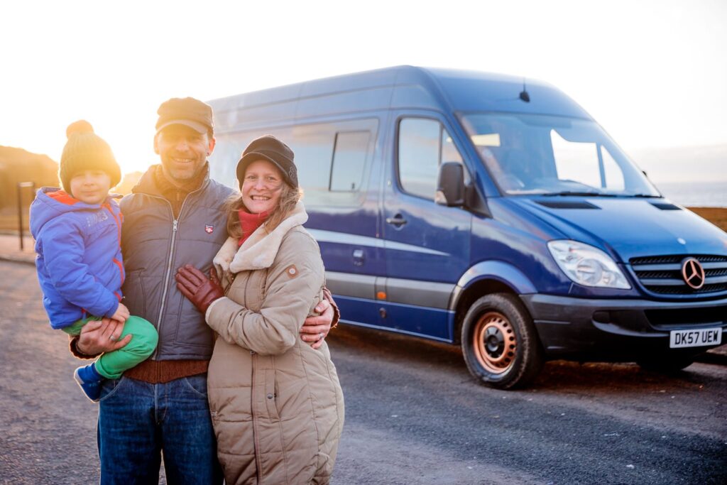 A family of three stands in front of a blue Mercedes-Benz Sprinter van. An adult man with a beard holds a young child dressed in a blue coat and red beanie. A woman in a beige coat and black hat stands to their right. They all smile and stand close together with a sunset in the background.