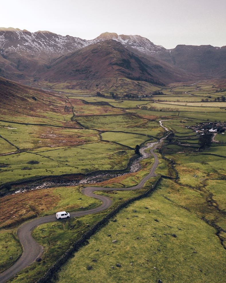 A winding road curves through a lush, green valley with a white car driving along it. Patches of brown and green fields, divided by low stone walls, spread across the landscape. A narrow river follows the road's direction. Snow-capped mountains rise in the distance under a pale sky.