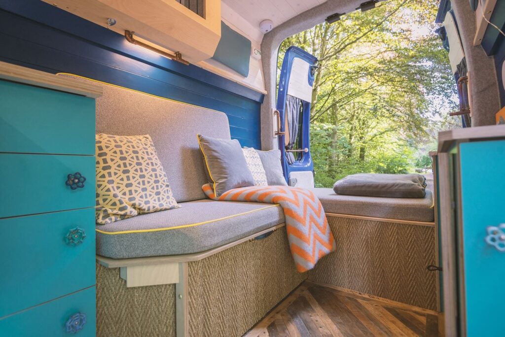 Interior of a converted camper van featuring a cozy seating area with gray cushions, patterned and plain throw pillows, and an orange chevron blanket. Teal cabinetry with flower-shaped knobs adds a pop of color. The back door is open, revealing a lush, green forest outside.