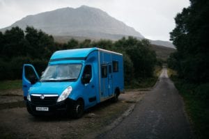 A blue selfbuilt campervan parked on the roadside with mountains in the background