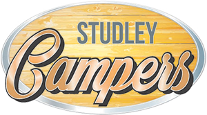 Logo of Studley Campers featuring an oval shape with a wood plank texture background. The word "Studley" is written in uppercase, blue-gray letters at the top, and "Campers" is written in large, cursive, metallic-looking letters in the center. The oval is outlined in gray.