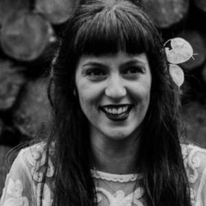 A black and white portrait of a smiling woman with long, dark hair adorned with a flower on the right side. She has bangs and is wearing a patterned lace top. The background is blurred but appears to be a stack of wood or logs.