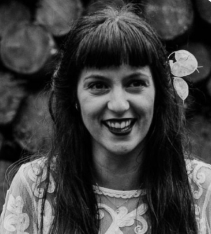 A black and white portrait of a smiling woman with long, dark hair adorned with a flower on the right side. She has bangs and is wearing a patterned lace top. The background is blurred but appears to be a stack of wood or logs.