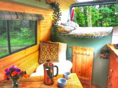 A cozy camper van interior features a small wooden dining table with a French press and two mugs, and a cushioned seating area with patterned pillows and a throw blanket. A raised bed is in the background near a window with green trees outside. A vase of colorful flowers sits on the table.