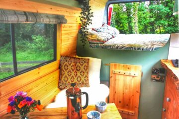 A cozy camper van interior features a small wooden dining table with a French press and two mugs, and a cushioned seating area with patterned pillows and a throw blanket. A raised bed is in the background near a window with green trees outside. A vase of colorful flowers sits on the table.