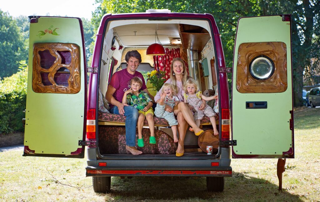 A family of five sits at the open back of a converted camper van. The parents sit in the middle, each holding a toddler, one boy and one girl, while an older boy stands in front of them. The van's interior is decorated with wood accents, and a red hanging lamp is visible inside. Trees are in the background.