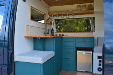 The interior of a converted camper van is shown. It features teal lower cabinets, a small refrigerator, a wooden countertop, a sink with a tall faucet, and glass jars stored above. A cushioned bench is on the left, and the van door reveals an outside view of greenery.