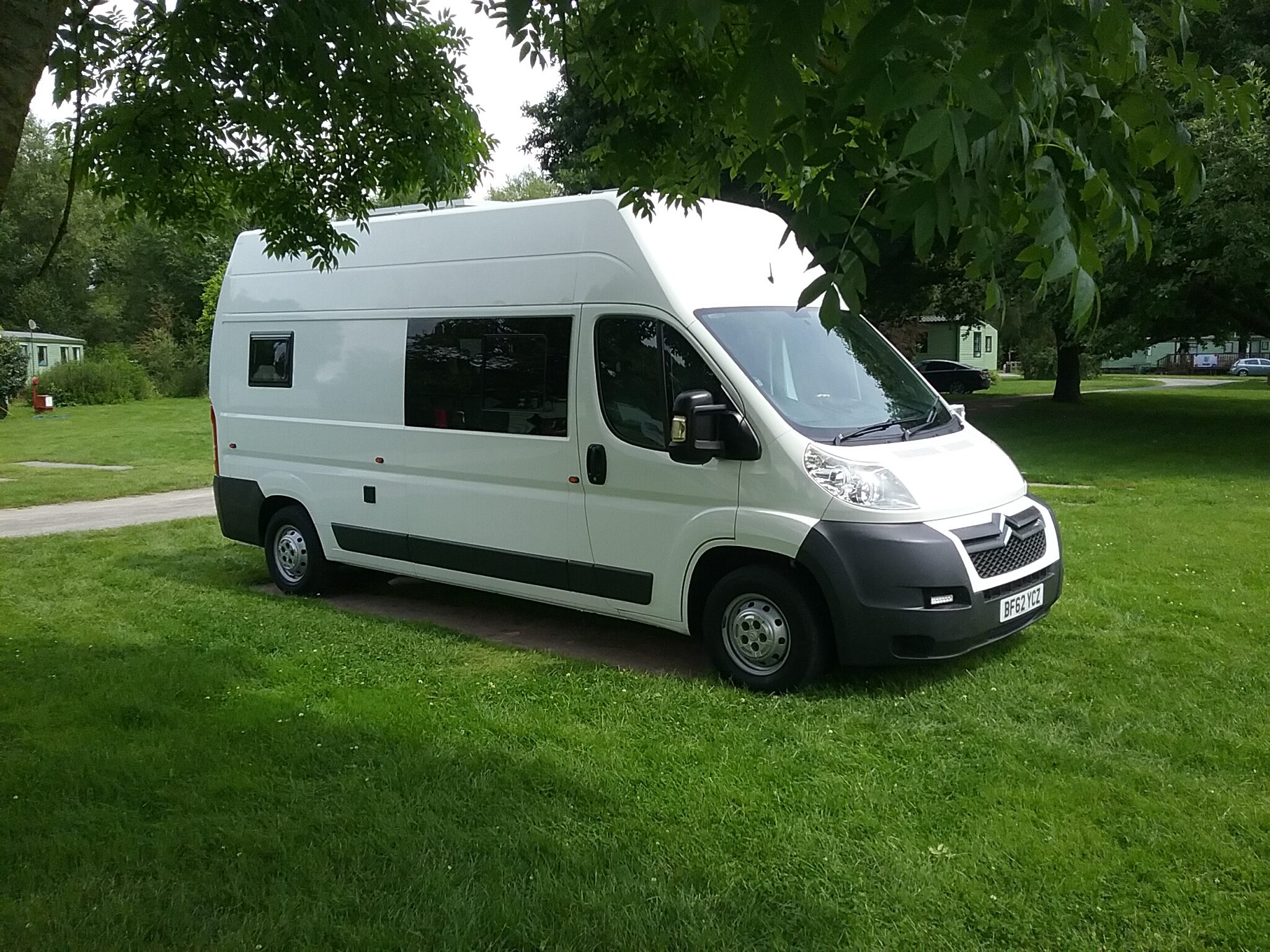 A white campervan is parked on a grassy area surrounded by trees. The van features a high roof, black side trim, and a large window on the side. Sunlight filters through the tree canopy, casting dappled shadows on the vehicle. In the background, other RVs are visible among the greenery.