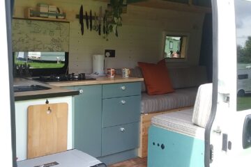 The interior of a camper van is shown with an open door. Inside, there's a small kitchenette with blue cabinets, a countertop with utensils hanging above, and a compact fridge adorned with stickers. A striped couch with orange and beige cushions sits beside a window. Some potted plants hang from the ceiling.