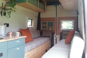 The interior of a converted camper van features two cushioned benches with orange pillows, a small kitchen area with cupboards and a countertop, hanging plants, and a window on each side. The walls and ceiling are finished in light wood. There is also a small set of shelves on the back wall.