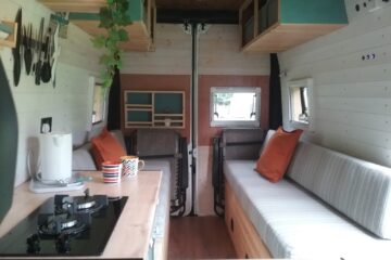 Interior of a cozy van conversion featuring a kitchenette with a stove on the left, a striped sofa with orange cushions on the right, and small windows on both sides allowing natural light. The walls are lined with light-colored wooden panels, and there are hanging plants and shelves for storage.