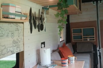 A cozy camper van interior with wooden paneling. It features a small kitchen area with a gas stove, kettle, and cutting board. Above, there are shelves with utensils and plants. The living space includes a cushioned bench with pillows and a map of Italy on the wall. Two mugs sit on the counter.