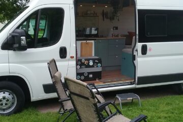 A white camper van with its side door open reveals a compact interior. Four folding chairs are set up on the grassy area outside. Inside the van are visible kitchen elements including a counter, sink, and cupboards. The scene depicts a cozy and organized mobile living space.