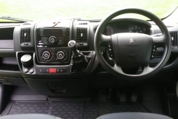 The image shows the interior of a Citroën vehicle's cockpit from the driver's perspective. It features a multifunction steering wheel, a central control panel with knobs and buttons for air conditioning and media, a gear stick, cup holders, and an instrument cluster behind the steering wheel.