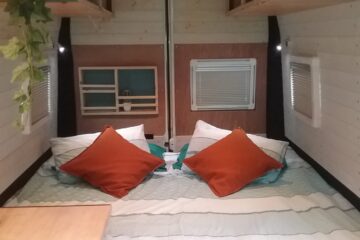 A cozy camper van interior featuring a neatly made bed with white and teal bedding. The bed is adorned with two white pillows and two orange cushions. Above the bed are teal overhead storage compartments. There is a small plant hanging from the upper left corner, and a wooden countertop is partially visible on the left side.