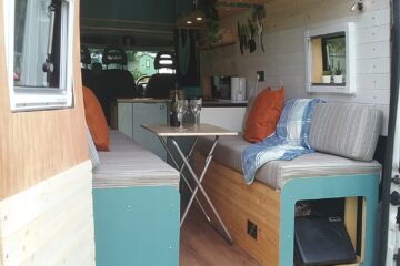 A converted camper van with the rear doors open reveals a cozy interior. The space includes two cushioned bench seats with orange throw pillows, a folding table in between, and overhead storage cabinets. The wooden paneling and plants add a homey touch. A front cab area is visible beyond the living space.