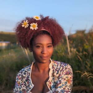 A woman with curly reddish-brown hair adorned with three white daisies is standing outdoors. She is wearing a floral-patterned top and is looking directly at the camera with a gentle smile. The background features blurred greenery and hints of a blue sky, suggesting a natural setting.