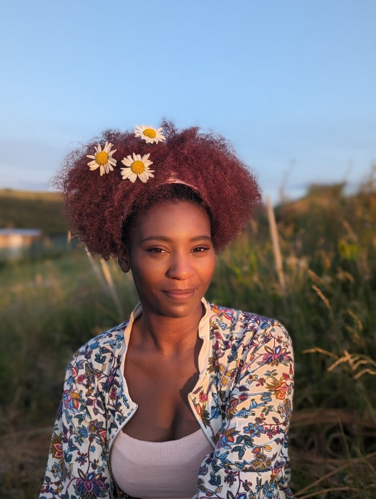 A woman with curly reddish-brown hair adorned with three white daisies is standing outdoors. She is wearing a floral-patterned top and is looking directly at the camera with a gentle smile. The background features blurred greenery and hints of a blue sky, suggesting a natural setting.