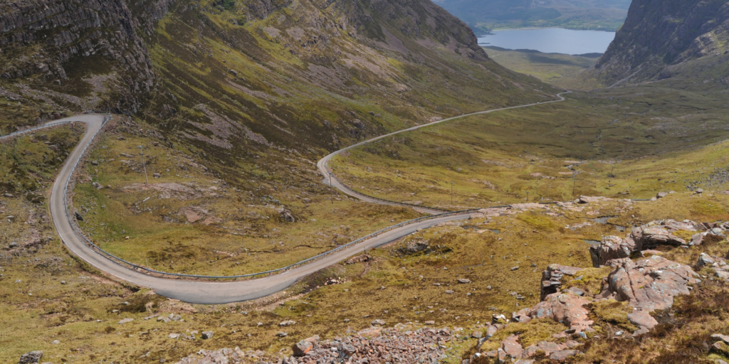 The Guide takes you along a winding path through a lush green valley, flanked by steep, rocky fells. The narrow route twists sharply as it climbs the terrain. In the backdrop, you can see a serene lake nestled among the fells beneath a partly cloudy sky. This pristine and natural scene exudes tranquillity.