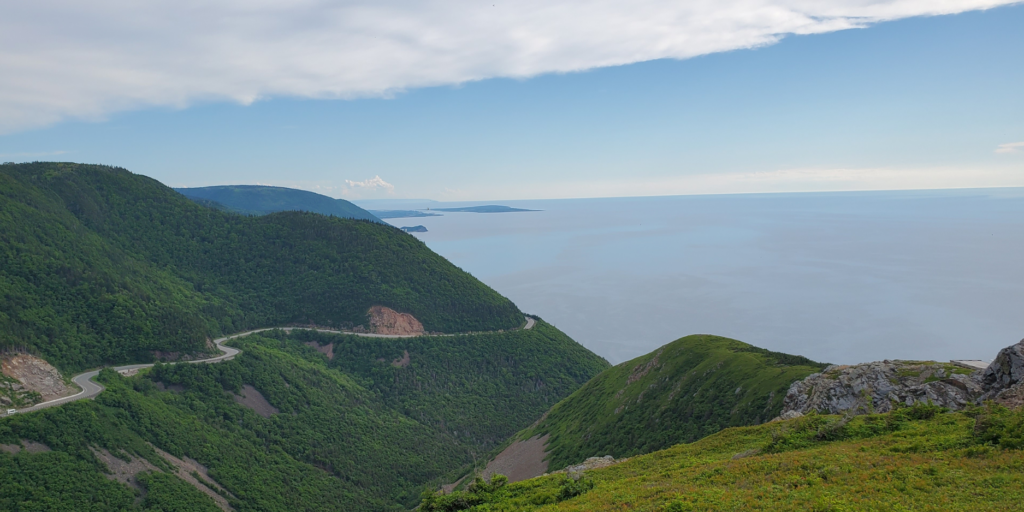 A winding road meanders through lush green hills and dense forest along a coastal mountain range, leading to a calm blue sea, reminiscent of scenes from an NC500 Guide. The sky is partially cloudy, with the horizon stretching out. To the right of the image is a rocky outcrop with green vegetation extending down towards the ocean