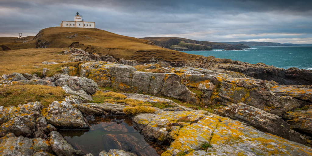 A windswept coastal scene shows a white lighthouse perched atop a hill. The rocky terrain in the foreground features patches of light green and yellow moss. On the right, the sea is rough with waves crashing, while heavy clouds dominate the overcast sky above. In the distance, brown hills and cliffs stretch into the horizon.