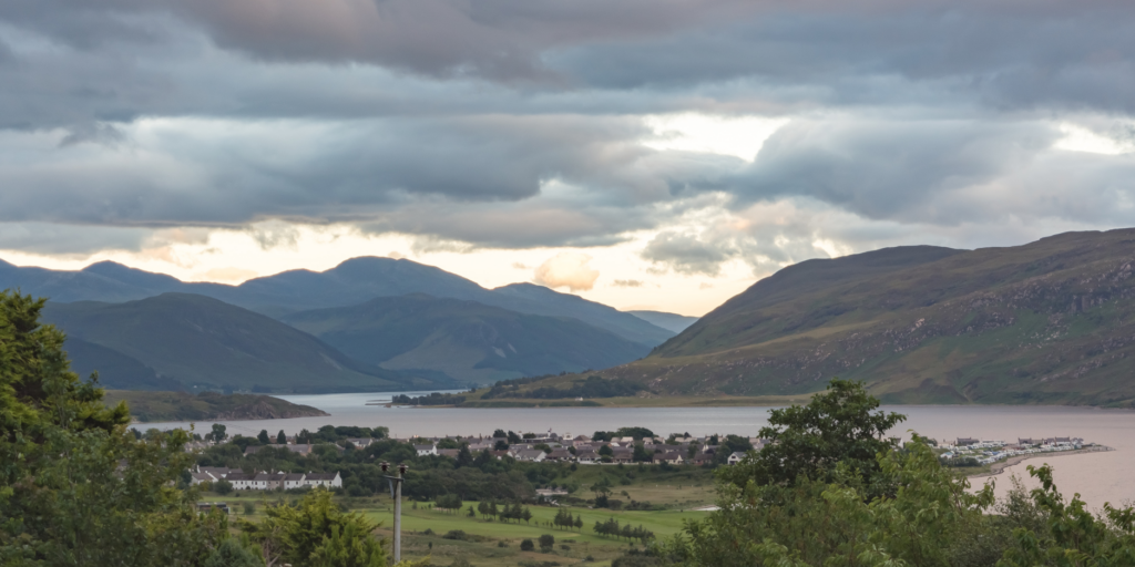 A serene landscape photo showcasing a picturesque village by a tranquil lake, surrounded by rolling green hills and mountains beneath a cloudy sky at dusk. Tthe village has scattered houses, lush vegetation encircles the lake, and mountains provide a stunning backdrop.