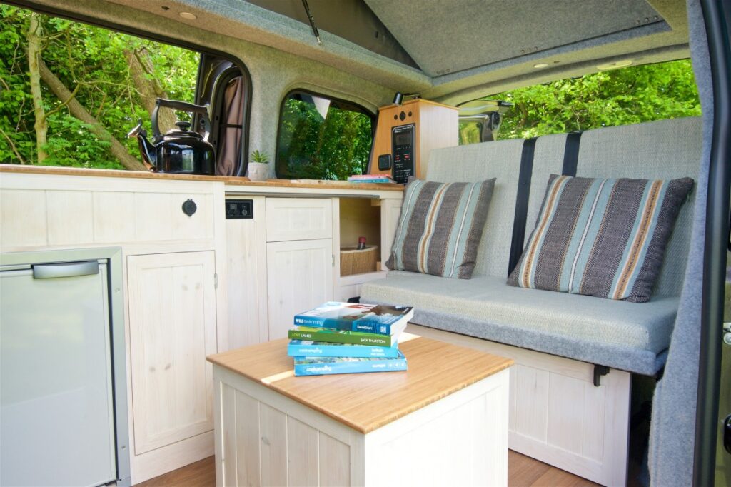 The interior of a camper van features a compact kitchen area with a kettle and microwave, light-colored wooden cabinetry, and a mini-fridge. A cozy seating area with striped cushions is next to a small wooden table, topped with several books. Greenery is seen through an open window.