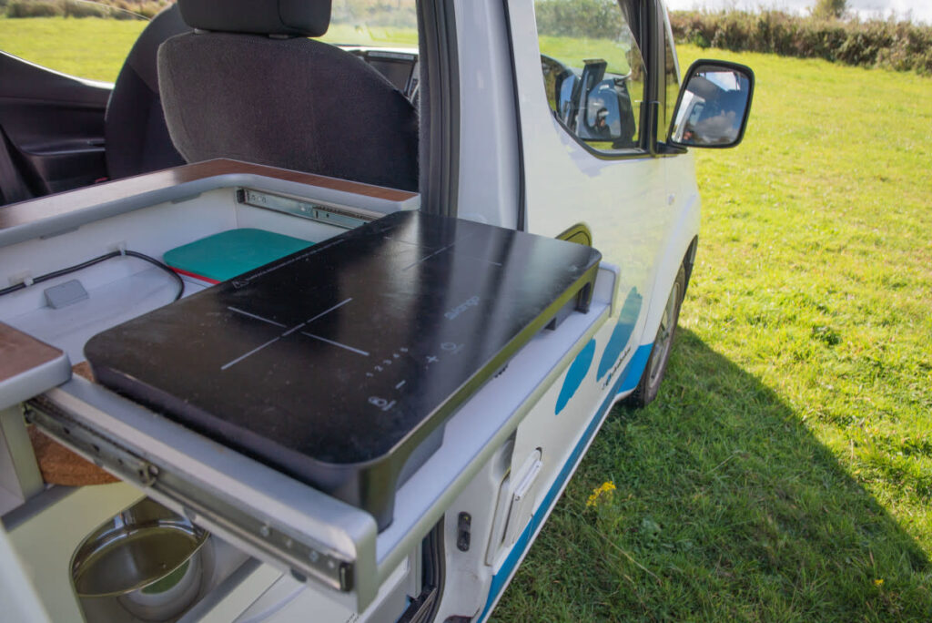 A compact camper van's kitchen setup with an extended slide-out counter featuring an induction cooktop. The van's side door is open, revealing a grassy field outside. There’s a metal bowl beneath the counter and a closed cabinet with kitchen supplies inside the van.