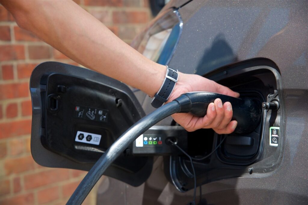 Close-up image of a person’s hand plugging a black charging cable into the port of an electric vehicle. The vehicle is positioned against a brick wall in the background. The person's wristwatch is visible, and the car's charging port cover is open, showing various labels and indicators.