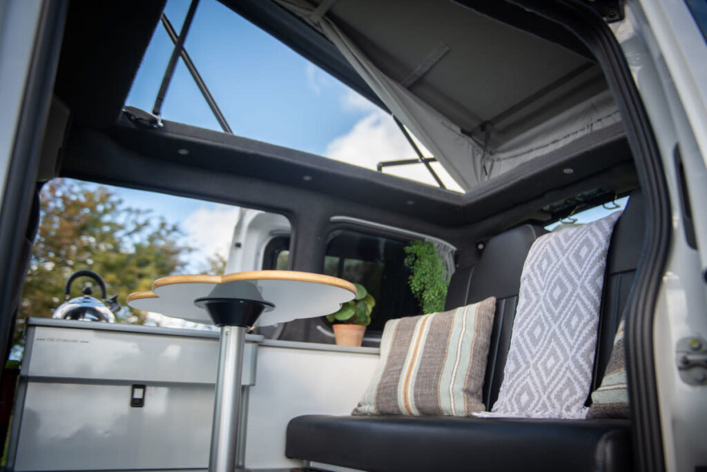 Interior view of a camper van with a pop-up roof. It features a small table with a light wood top on a single metal leg. The seating area has cushions in striped, geometric, and solid patterns. A teapot and a small plant sit on the kitchenette counter, with greenery visible through the windows.