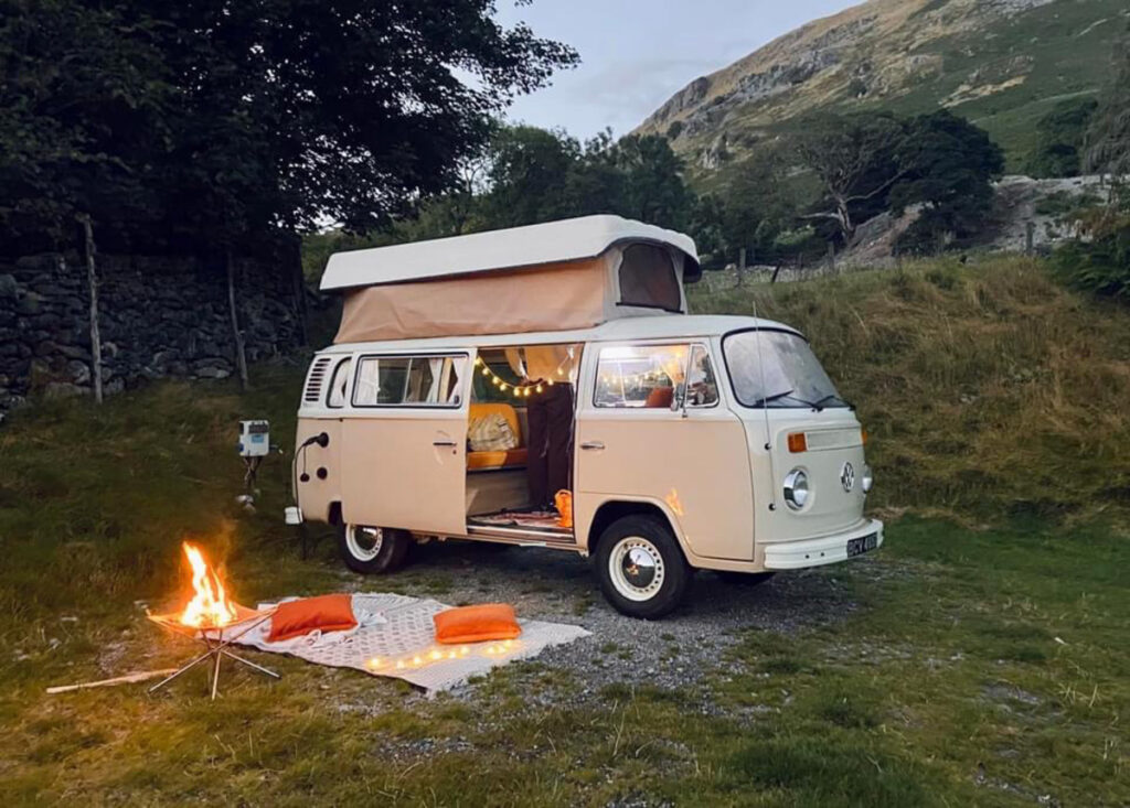 A cream-colored, vintage camper van with an elevated pop-top roof is parked on a grassy area near a stone wall and trees. The van is decorated with string lights and has an open door showing a cozy interior. Outdoors, a small campfire burns next to a patterned mat and two orange cushions.