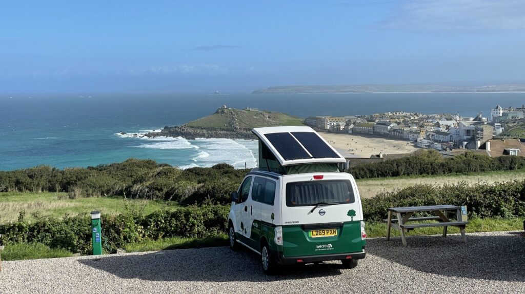 A white and green camper van is parked on a gravel area overlooking a coastal landscape. The van's roof is equipped with a solar panel. In the distance, waves crash against the shore, with a small town and a peninsula featuring a prominent hill on the horizon. A picnic table is nearby.