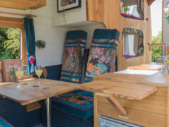 Interior of a cozy camper van with colorful patterned cushions, a wooden fold-out table, and two glasses of wine on the table. A small vase with flowers adds charm to the setting. The right side features a wooden structure with windows that let in natural light, enhancing the warm atmosphere.