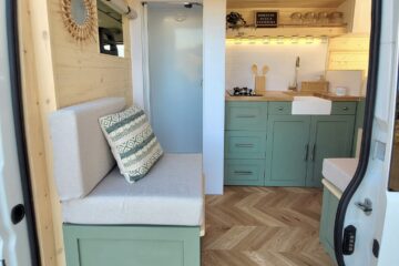 Interior of a converted camper van showcases a cozy living space featuring a built-in cushioned bench with decorative pillows on the left. The right side holds a green kitchenette with drawers, a farmhouse sink, and floating shelves, all under warm LED lighting. The floor is herringbone-patterned wood.