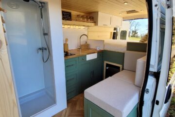 Interior of a converted van showcasing a cozy living space. On the left, there's a white shower with a glass door. The middle has a kitchen area with green cabinets, a sink, and wooden countertop. On the right, a seating area with beige cushions. A bed is visible above the seating area.