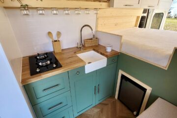 A cozy RV kitchen with a wooden countertop, green cabinets, a small white sink, and a black two-burner stove. Above the sink are clear jars hanging on hooks. There is a mini-fridge below the counter and a wooden shelf above holding utensils and a small basket. The area is well-lit by natural light.