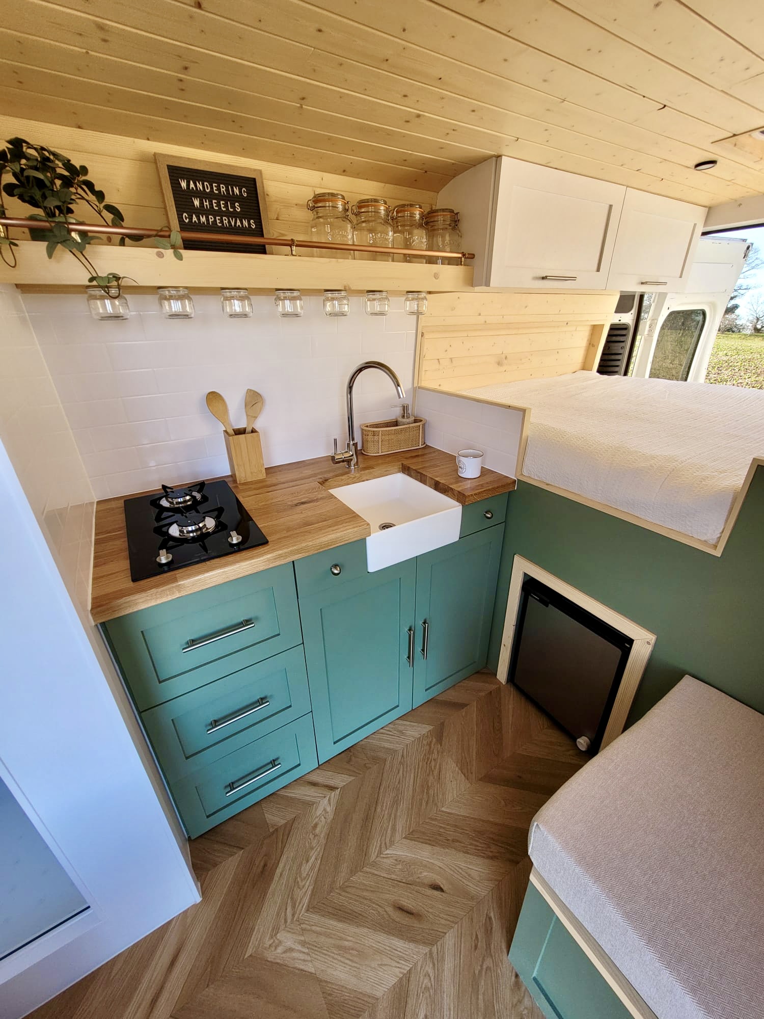 A cozy RV kitchen with a wooden countertop, green cabinets, a small white sink, and a black two-burner stove. Above the sink are clear jars hanging on hooks. There is a mini-fridge below the counter and a wooden shelf above holding utensils and a small basket. The area is well-lit by natural light.