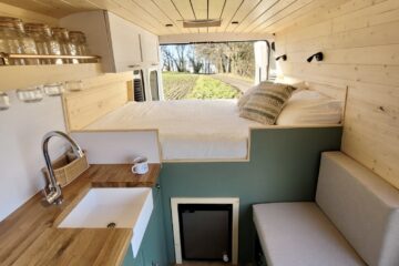 Interior of a minimalist camper van featuring a cozy bed elevated above a bench seat, adjacent to a small kitchenette. The kitchen area includes a white sink, wooden countertops, and glass jars on a shelf. The walls and ceiling are lined with light wooden paneling, creating a warm, inviting atmosphere.