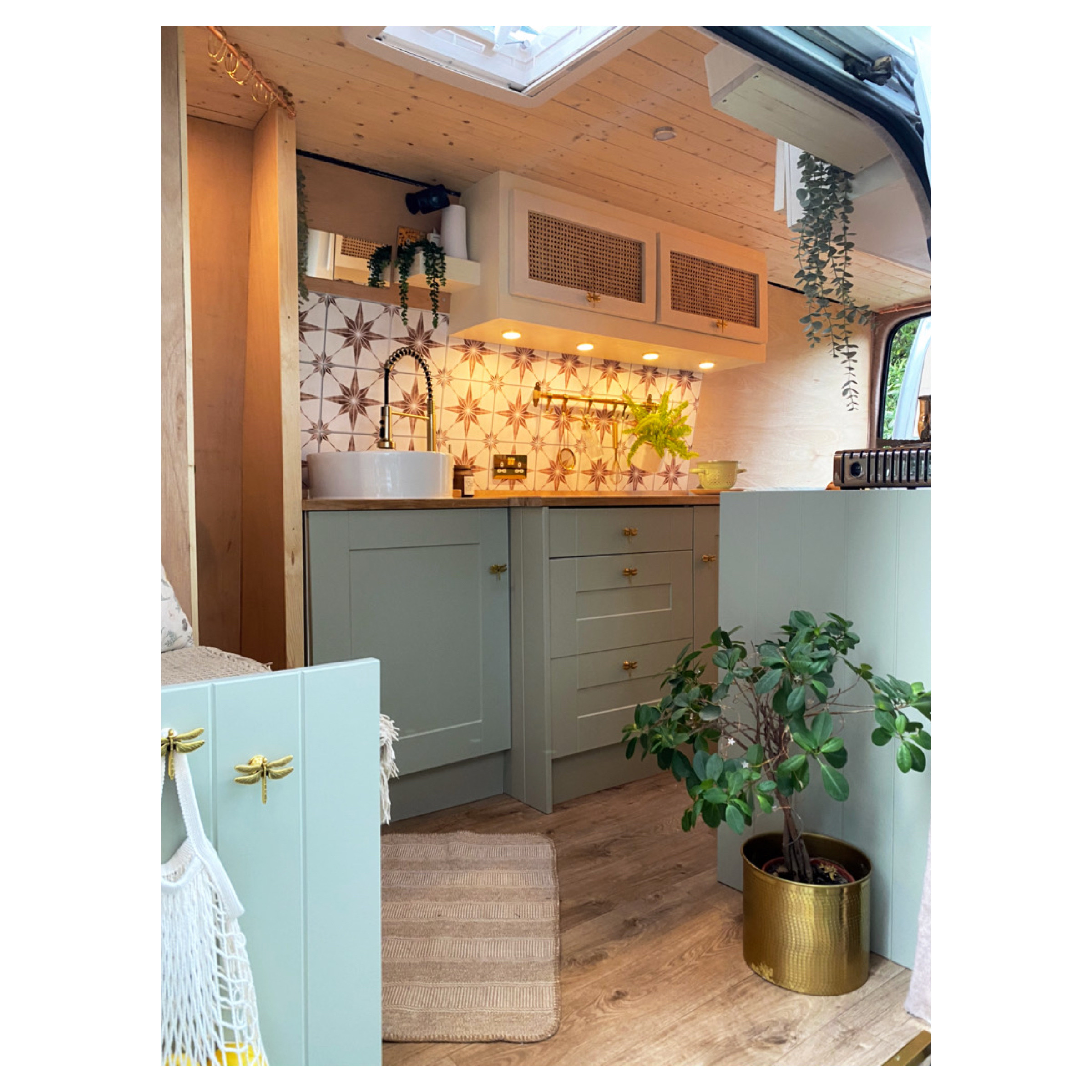 A cozy van interior with a kitchenette featuring mint green cabinets and drawers, a white round sink, and a star-patterned backsplash. Potted plants adorn the space including one in a gold pot. A hammock chair is partially visible to the left, and a skylight provides natural lighting.