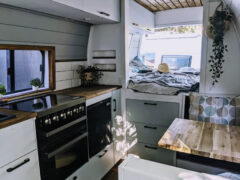 A compact, modern kitchen inside a van, featuring a stove, oven, sink, and wooden countertops. A cozy bed with pillows and blankets is at the rear, near a window letting in light. There is a small dining area with a cushioned bench and wooden table. Plants and decorations accent the space.