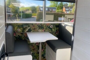 A small dining nook in a compact space features a white table with a single metal leg, flanked by two gray cushioned benches. The back wall is adorned with colorful floral wallpaper. A large window offers a view of suburban houses and greenery, while potted plants sit on a shelf above.