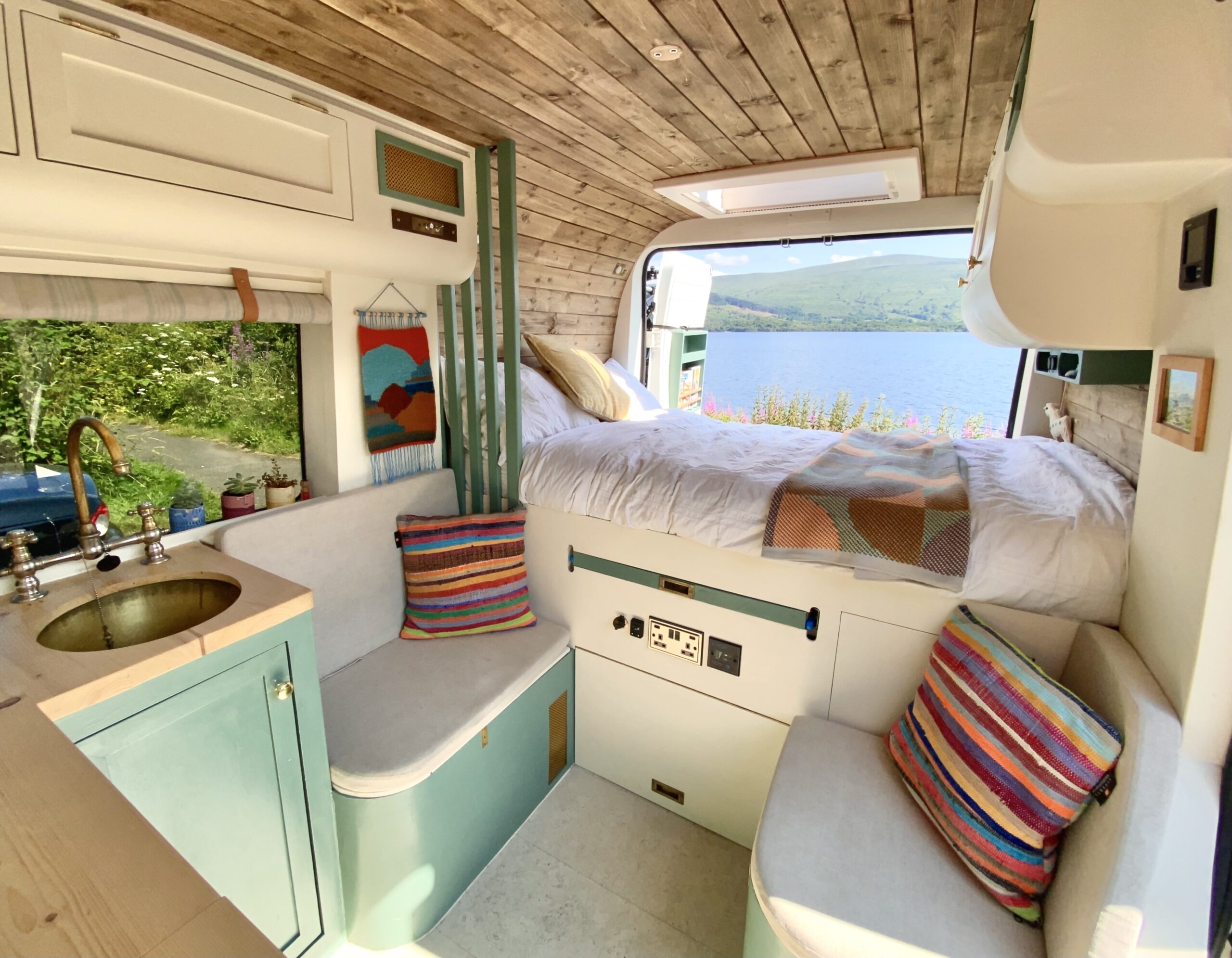 Interior of a cozy camper van with a wooden ceiling and white-paneled walls. A kitchenette with a small sink and stove occupies the left side, while a bed with white bedding is set up at the rear, near a large window showing a scenic lakeside view. Two cushioned seats with colorful striped pillows are near the kitchen.