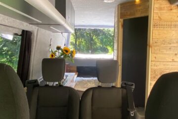 Interior of a camper van showing a cozy setup. Front seats face a small table. The rear features a bed with white bedding adorned with a vase of sunflowers. Walls are wood-paneled. Windows provide natural light, revealing lush greenery outside. A ceiling vent and storage cabinets are visible.