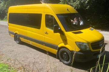 A bright yellow Mercedes-Benz van is parked on a sunlit path beside a dense green forest. The van is large and has tinted windows. The driver’s side window is rolled down, revealing two people inside. The path is unpaved and surrounded by grass and tall plants in the foreground.