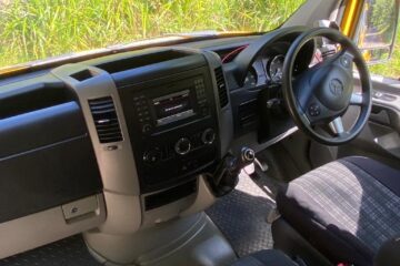 Interior of a vehicle showcasing the driver’s area. The dashboard features a central control panel with a screen and various knobs and buttons. The steering wheel, with the Mercedes-Benz logo, is on the right side, indicating a right-hand drive. The seats are fabric, in a checked pattern.