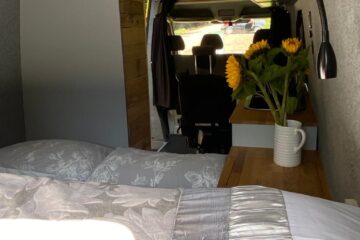 Interior of a cozy camper van with a neatly made bed in the foreground. A wooden nightstand next to the bed holds a white vase of bright yellow sunflowers. The van's driving seats are visible in the background, and the side window allows sunlight to illuminate the space.