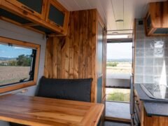 The interior of a camper van features a wooden table with bench seating on the left, wood-paneled walls, and overhead cabinets. The right side of the image shows a small kitchen area with a sink and tiled backsplash, and an open rear door reveals a scenic countryside view.