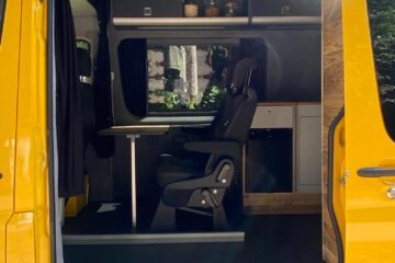 A bright yellow van with an open side door reveals a cozy interior setup. Inside, there is a swivel chair facing a foldable table and shelves with jars and decor items. The van has curtains for privacy, overhead lighting, and a rear kitchenette area. The background outside shows green foliage.