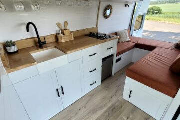 A camper van interior features a compact kitchen with white cabinets, a black faucet above a small white sink, and a wooden countertop. To the right, an L-shaped seating area with red-brown cushions surrounds a small round mirror on the wall, under a window showing a scenic outdoor view.