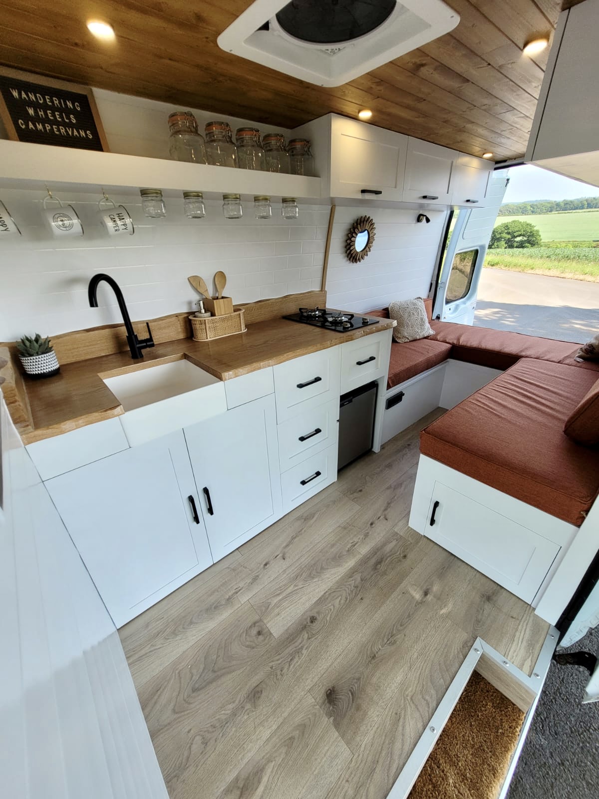 A camper van interior features a compact kitchen with white cabinets, a black faucet above a small white sink, and a wooden countertop. To the right, an L-shaped seating area with red-brown cushions surrounds a small round mirror on the wall, under a window showing a scenic outdoor view.